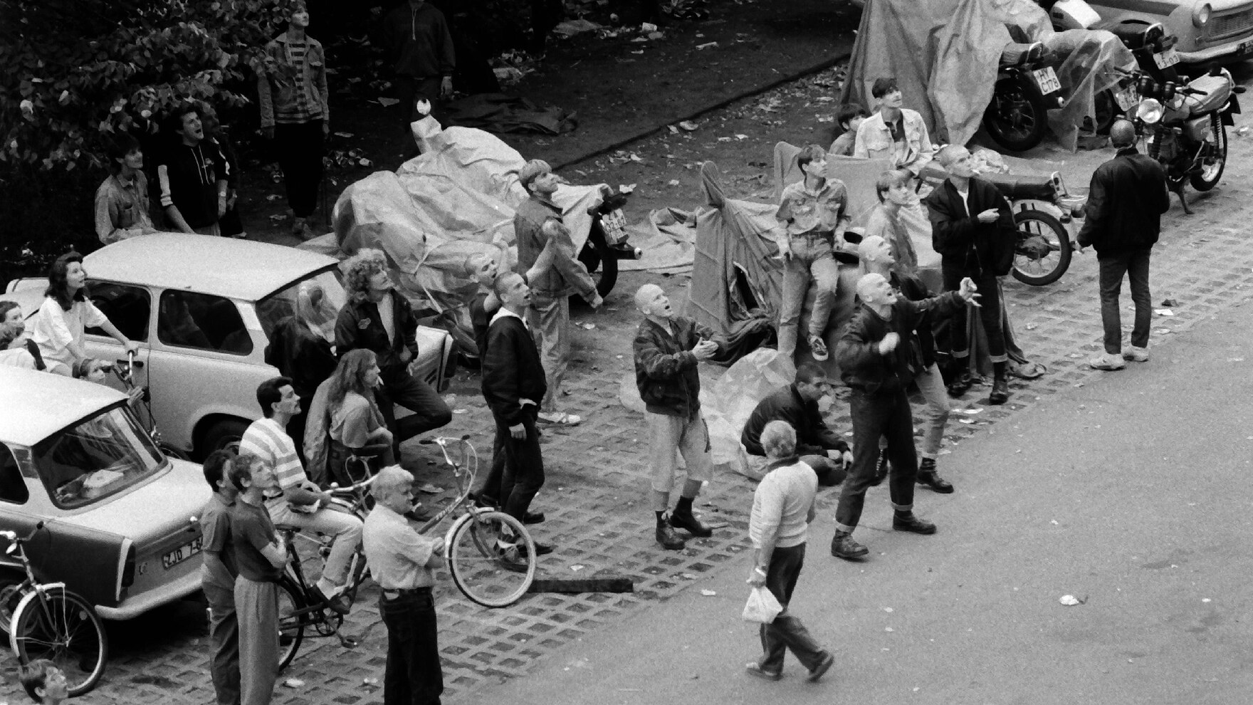 Eine Gruppe von Menschen hat sich im September 1991 auf einem Parkplatz vor einem Wohnheim versammelt, in dem Vertragsarbeiter leben. Bei einem Teil von ihnen handelt es sich erkennbar um Rechtsextreme, die aggressiv gestikulierend grölen.