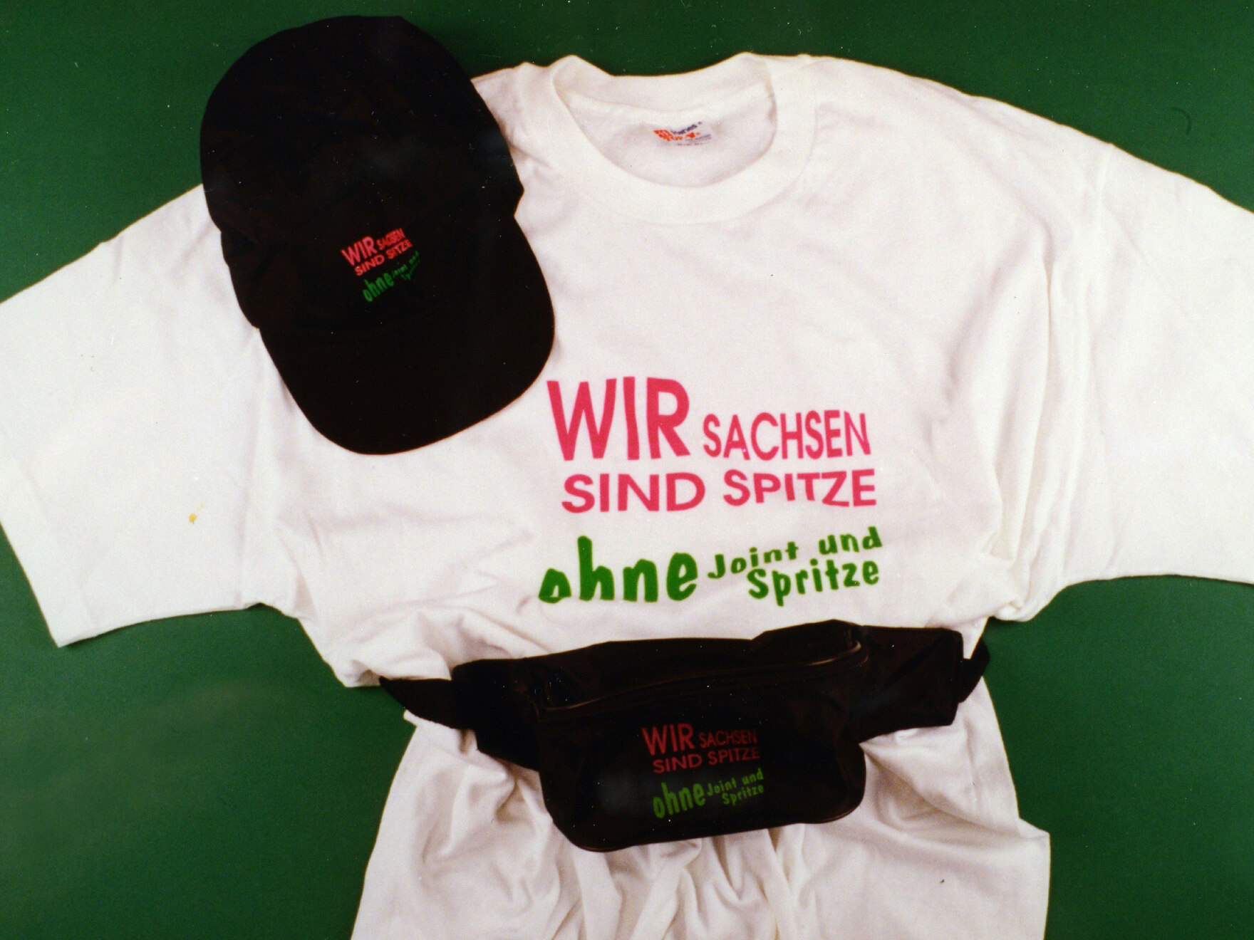 Im Rahmen der Präventionskampagne »Wir Sachsen sind Spitze ohne Joint und Spritze« wurden auch T-Shirts, Basecaps und Bauchtaschen mit dem Slogan bedruckt. Diese Kleidungsstücke sind vor einem grünen Hintergrund zu sehen.