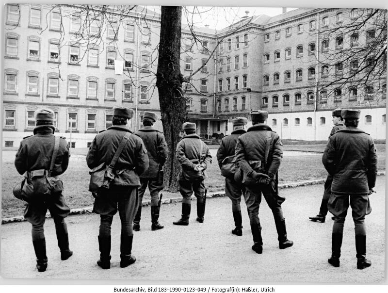 Sechs Polizisten stehen vor dem Strafvollzug Bautzen. Sie tragen Wintermützen und blicken auf die vergitterten Fenster der Vollzugseinrichtung. Gerade noch zu erkennen ist, dass sich hinter den Fenstern in der dritten Etage Menschen befinden.