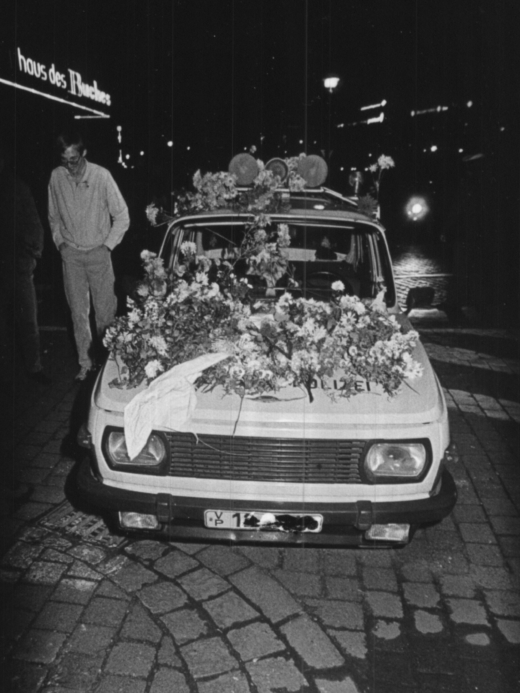 Ein abgeparkter Streifenwagen ist auf diesem Schwarz-Weiß-Foto auf der Motorhaube und dem Dach mit vielen Blumen bedeckt. Daneben ist ein Fußgänger zu erkennen.