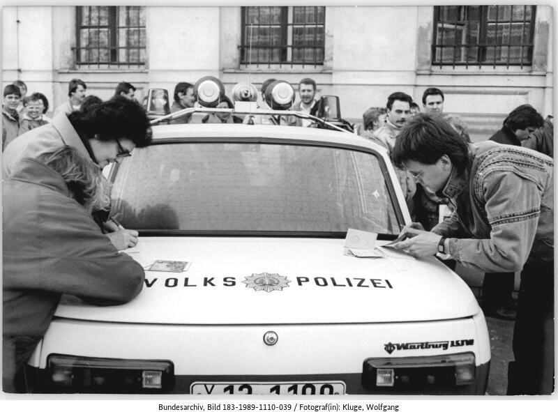 Auf der historischen Abbildung in schwarz-weiß ist ein Streifenwagen der Leipziger Volkspolizei zu sehen. Um ihn herum stehen mehrere Menschen. Drei Personen nutzen das Auto als Schreibunterlage.