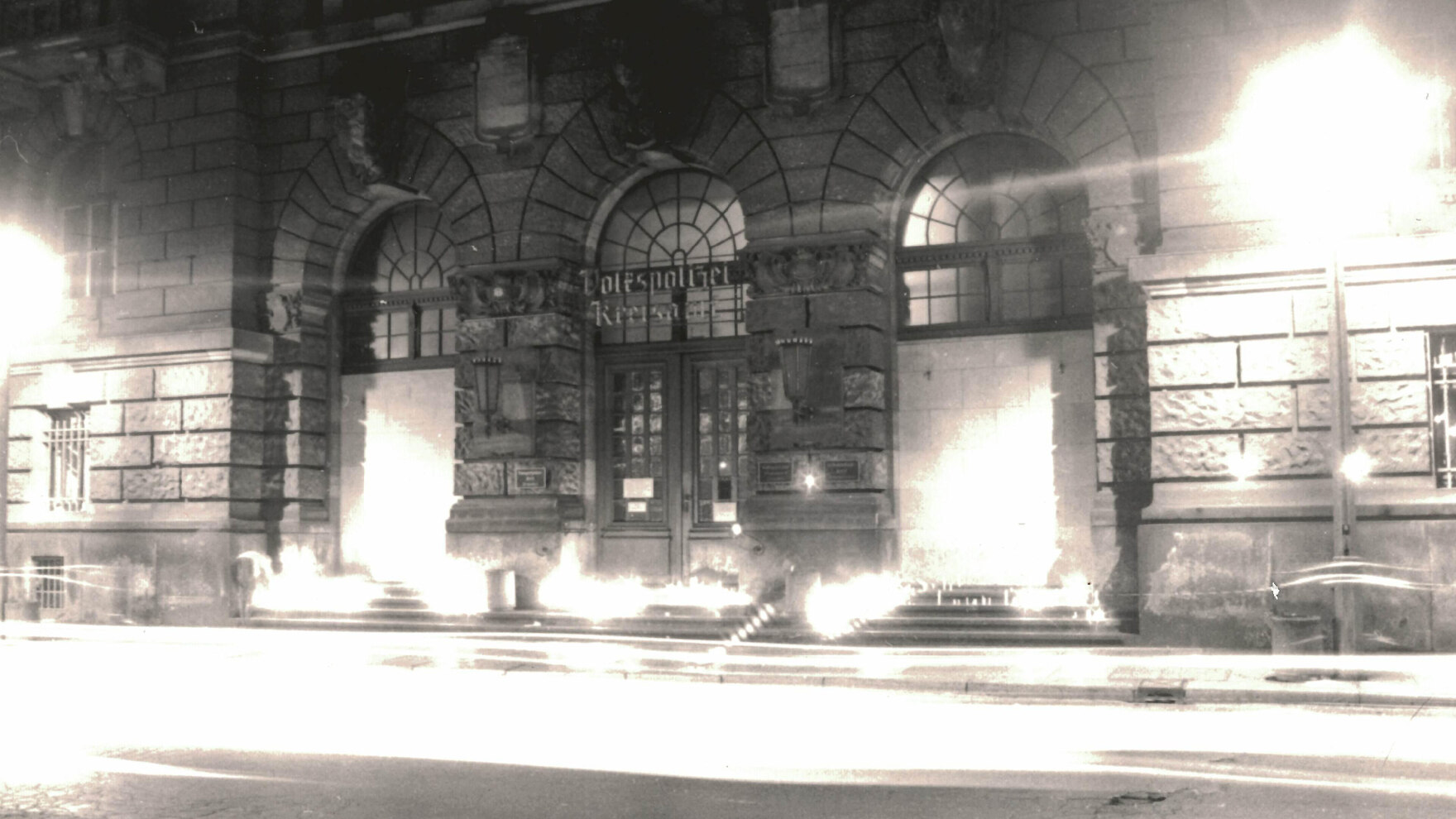 Das Foto in schwarz-weiß zeigt viele angezündete Kerzen auf den Stufen zu einem Gebäude, welches den Schriftzug »Volkspolizei Kreisamt« trägt. Das Bild ist an den hellen Stellen überstrahlt, die Kerzen an mehreren Stellen nicht einzeln zu erkennen.