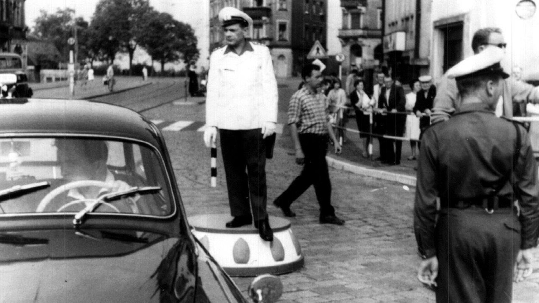 Auf dem Schwarz-Weiß-Foto ist ein Mann mit weißer Uniform eines Verkehrspolizisten auf einem Podest an einer Kreuzung zu sehen. Im Hintergrund sind viele Passanten, links vor dem Mann ein vorbeifahrendes Auto.