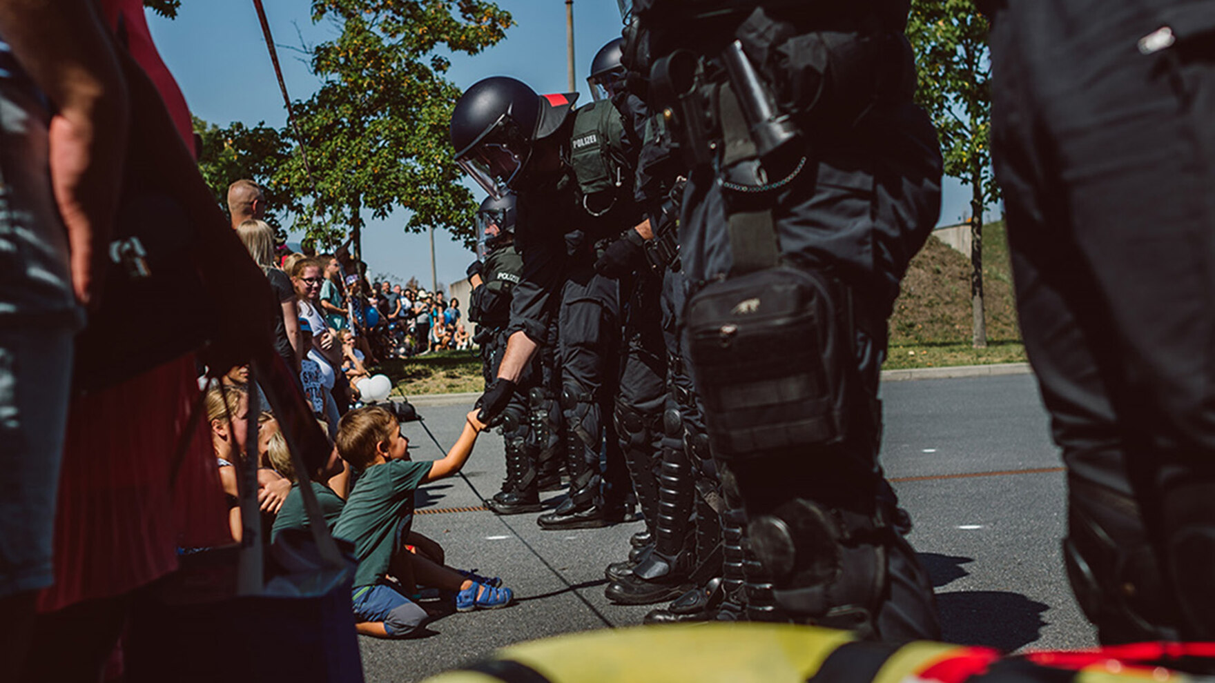 Einige Kinder sitzen und stehen mit weiteren Besuchern auf dem Fußboden vor uniformierten Bereitschaftspolizisten. Einer der Polizisten mit Helm beugt sich zu einem Kind herunter und schüttelt dem lächelnden Jungen die Hand.