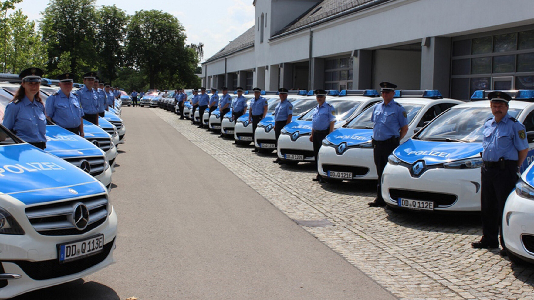 Polizistinnen und Polizisten in Uniform stehen vor einer Reihe Polizeiwagen auf einem Parkplatz. Es handelt sich um E-Autos.