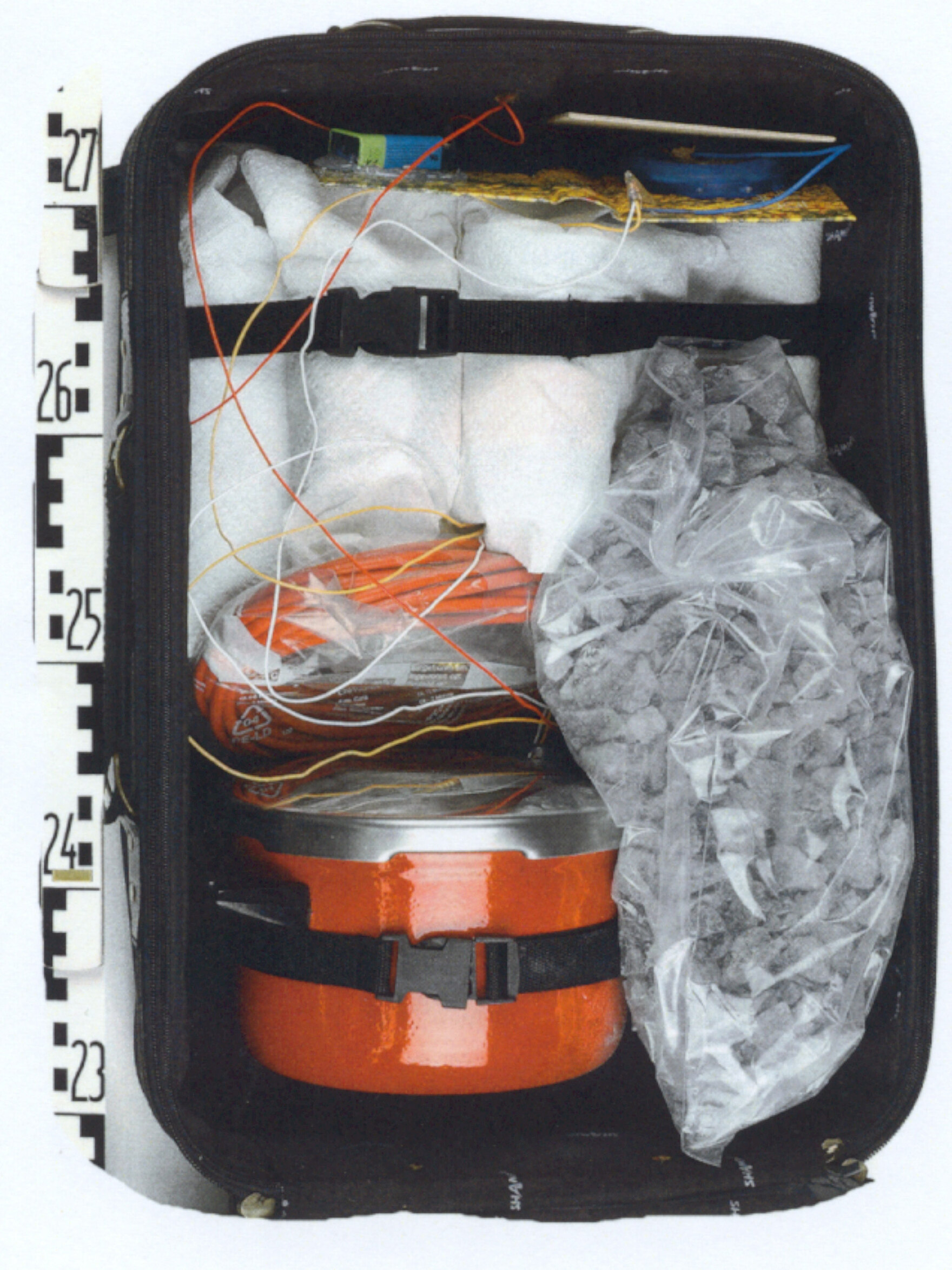 Dargestellt ist der Nachbau einer Kofferbombe. Dieser besteht unter anderem aus mehreren Kabeln, einem orangenen Kochtopf und einem Plastiksack, der mit Steinen gefüllt ist. Neben dem Nachbau liegt ein Lineal.