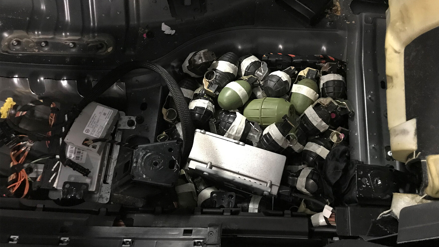 In dem Innenraum eines Autos unter der Verkleidung des Fahrersitzes liegen mehrere grüne und schwarze Handgranaten. Zu sehen ist außerdem ein Teil der Verkabelung des Wagens.