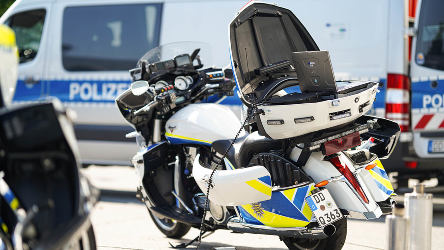 Ein Polizeimotorrad steht auf einem Parkplatz. Die Transportbox ist offen. Ein Laptop steht darin. Im Hintergrund ist ein größerer Polizeiwagen zu erkennen.