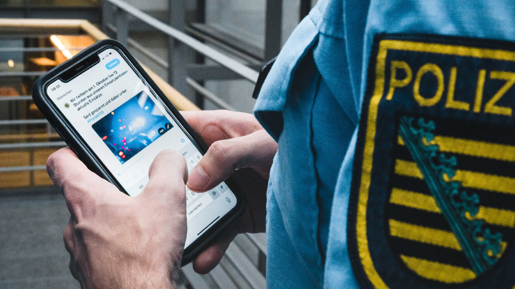 Ein Polizist in blauer Uniform hält ein Smartphone in der Hand. Er tippt gerade einen Tweet. Darin wird auf den Twitter-Marathon der Polizei Sachsen hingewiesen.