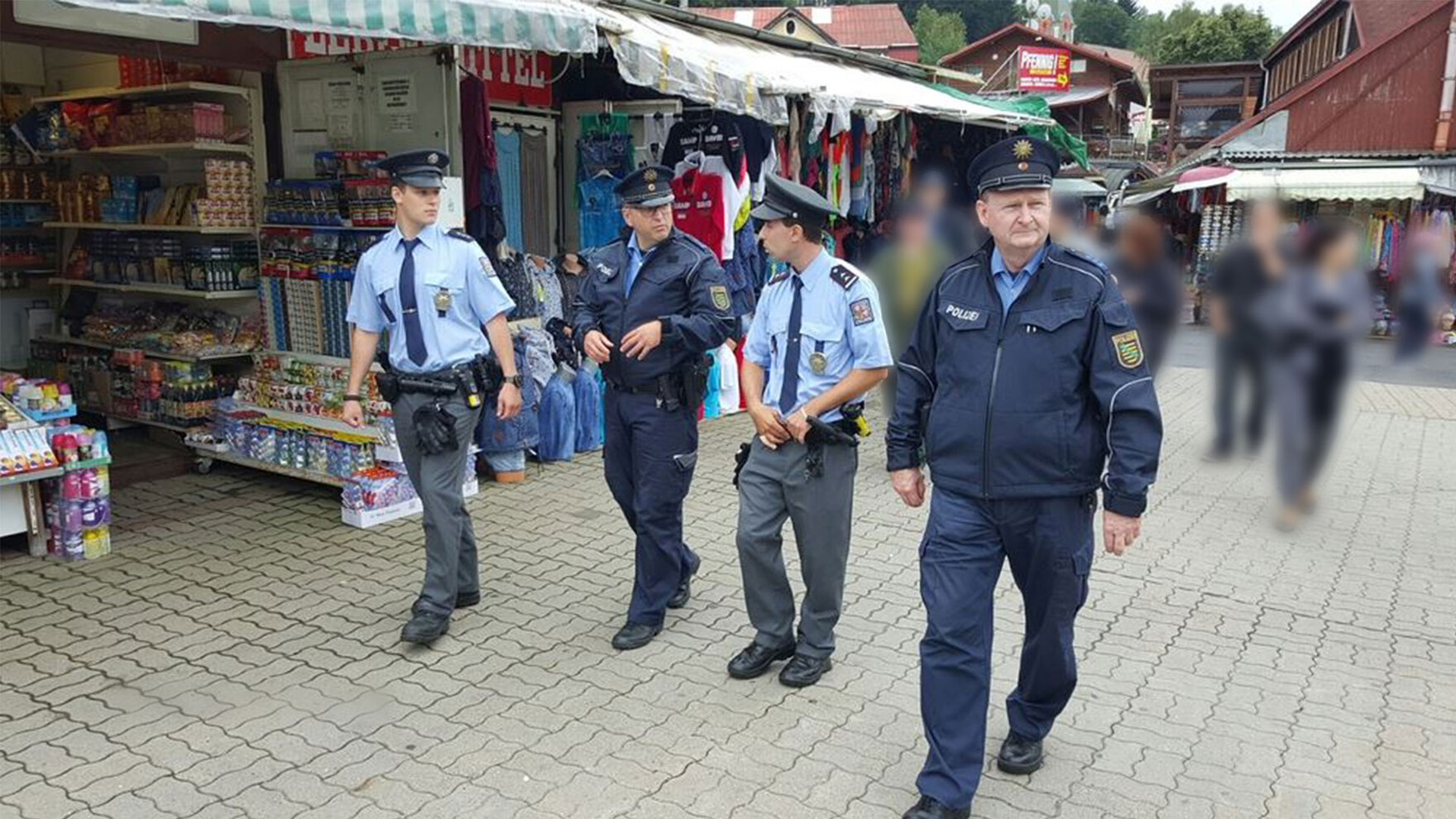 Vier Polizisten in Uniform – zwei aus Sachsen, zwei aus Tschechien – laufen über einen Marktplatz. Drei von ihnen besprechen sich dabei. Im Hintergrund sind andere Besucher des Marktes und verschiedene angebotene Waren zu sehen. 