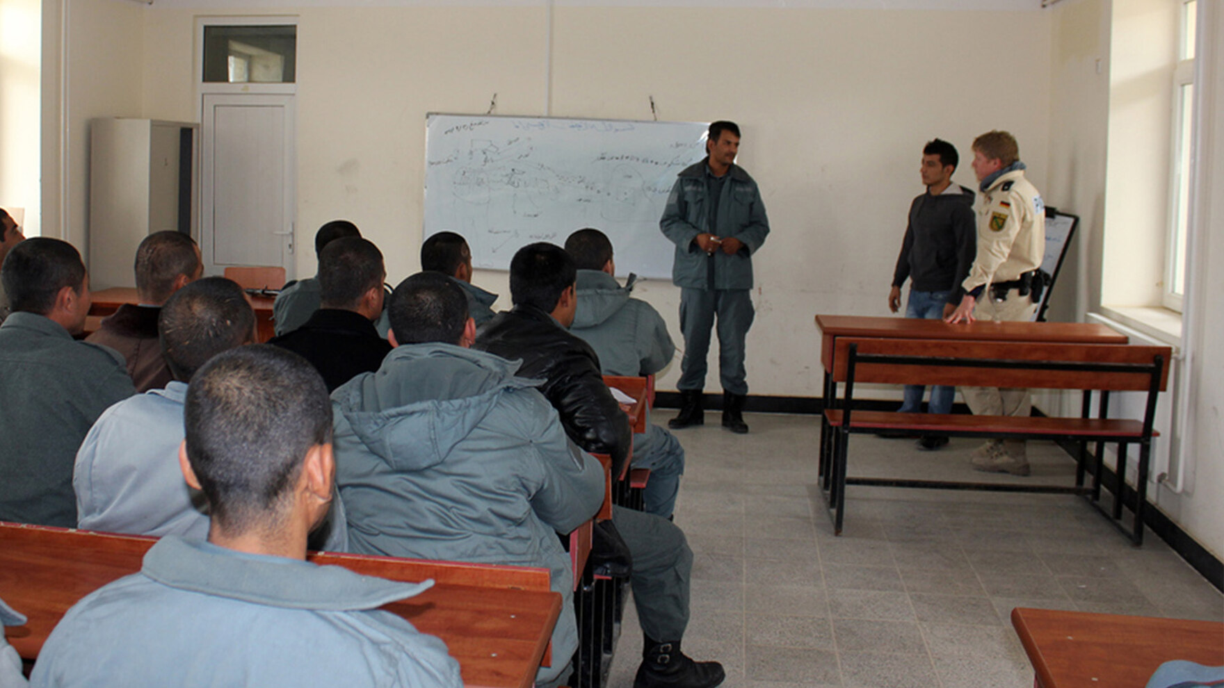 In einem Unterrichtsraum in Masar-e Scharif sitzen mehrere Männer an Bänken und schauen zu einer Gruppe von Personen vor einer Tafel. Auch ein sächsischer Polizist in heller Uniform steht vorne.