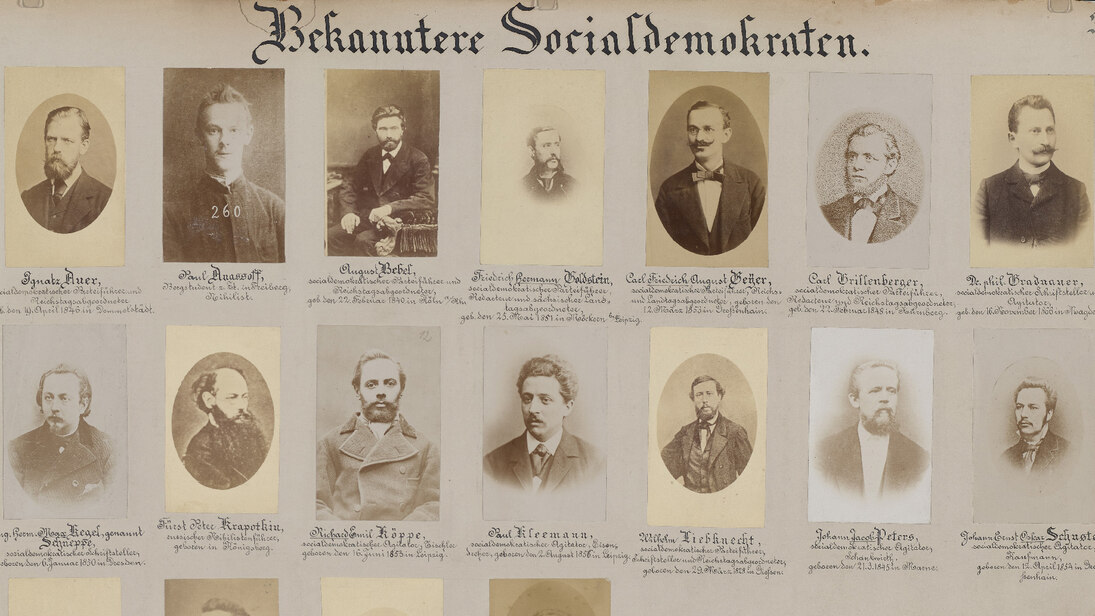 Die auf dieser historischen Lehrtafel aufgebrachten Fotografien zeigen unter der Überschrift »Bekanntere Socialdemokraten.« jeweils eine Person in Porträtaufnahme unterschiedlicher Gestaltung, darunter säuberlich jeweils den Namen und weiteren Text.
