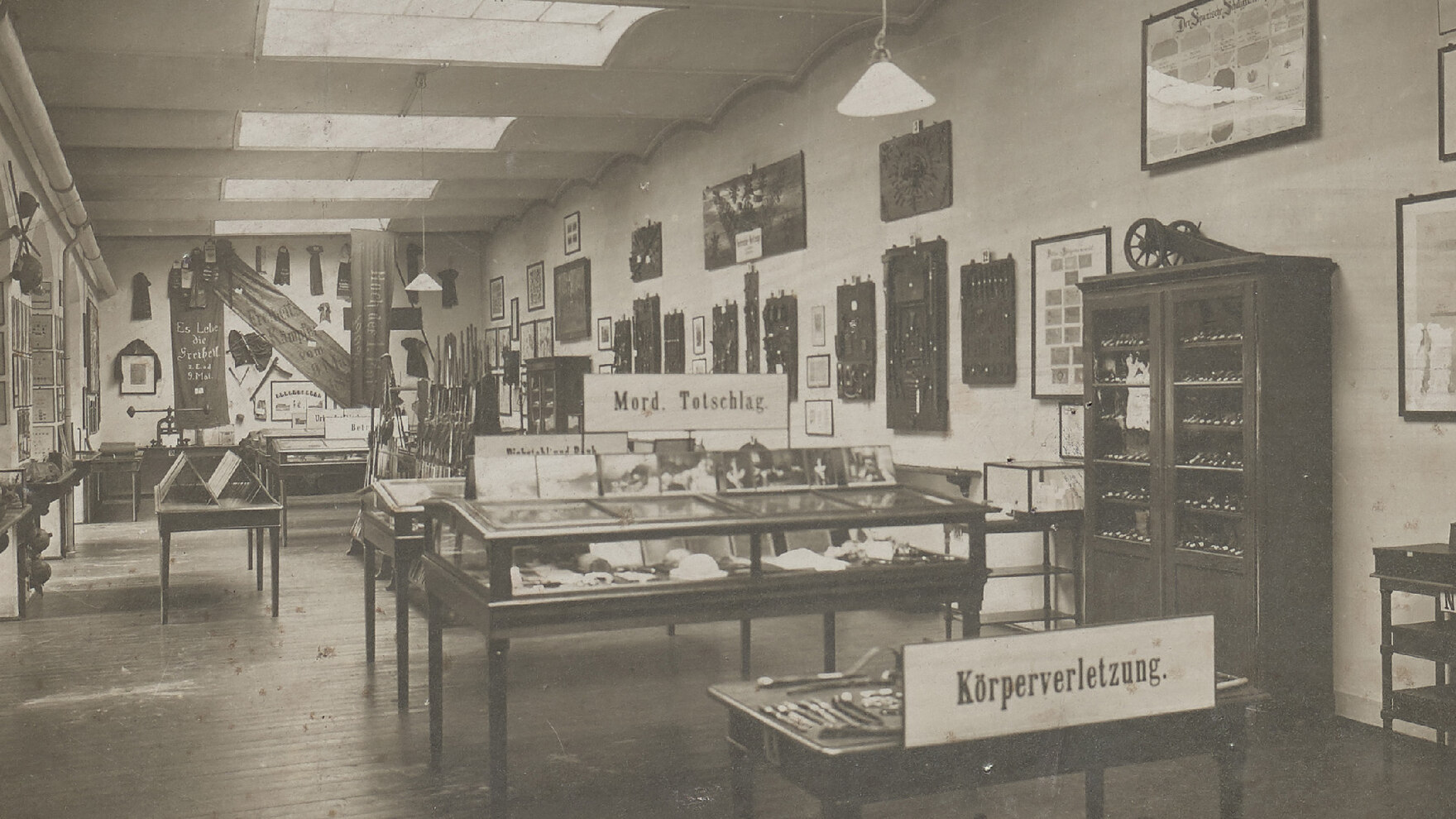 Das historische Bild zeigt den Blick in einen Raum des damaligen Kriminalmuseums. Auf einem Tisch steht ein Schild mit der Beschriftung »Körperverletzung«, dahinter ein Schild »Mord. Totschlag.«. Es sind sehr viele Ausstellungsstücke zu sehen.