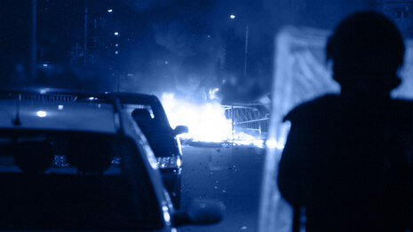 Das Foto zeigt rechts die Silhouette eines Bereitschaftspolizisten auf einer Straße neben Autos, vor ihm brennt etwas.