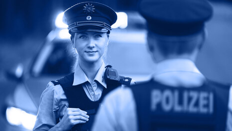 Eine junge Polizistin in Uniform schaut direkt in die Kamera. Hinter ihr ist ein Streifenwagen mit aktiviertem Blaulicht zu erkennen, vor ihr ein Kollege und auf seinem Rücken der Schriftzug »Polizei«.