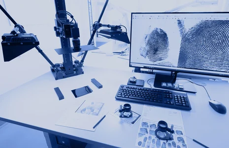 Ein daktyloskopischer Gutachterarbeitsplatz in einem sehr hellen Raum ist ausgestattet mit einer Kameraeinheit und einem Computer. Auf dem Computermonitor sind zwei Fingerabdrücke vergrößert zu sehen. Auf dem Tisch liegt ein Blatt mit Spuren und Lupe.